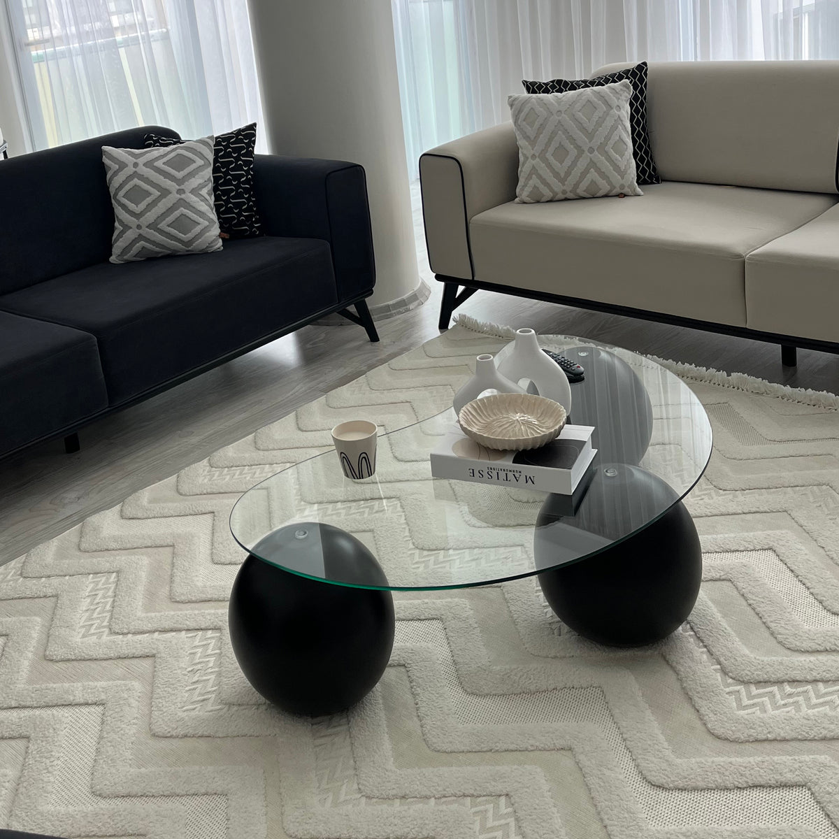 Glass coffee table with black ball legs on a white textured rug beside two sofas, front angle