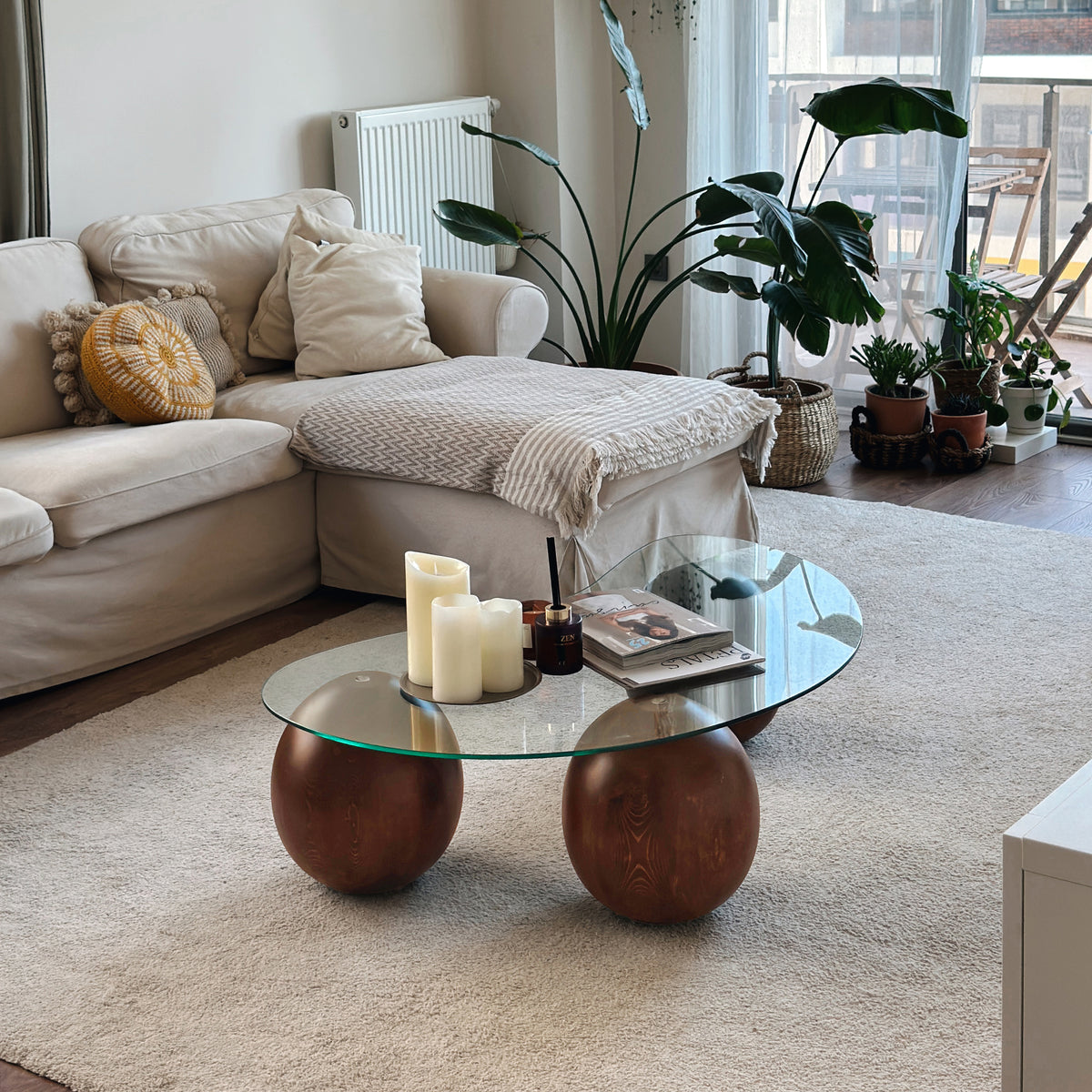 Glass coffee table with two wooden ball legs in front of a sofa and window, wide view