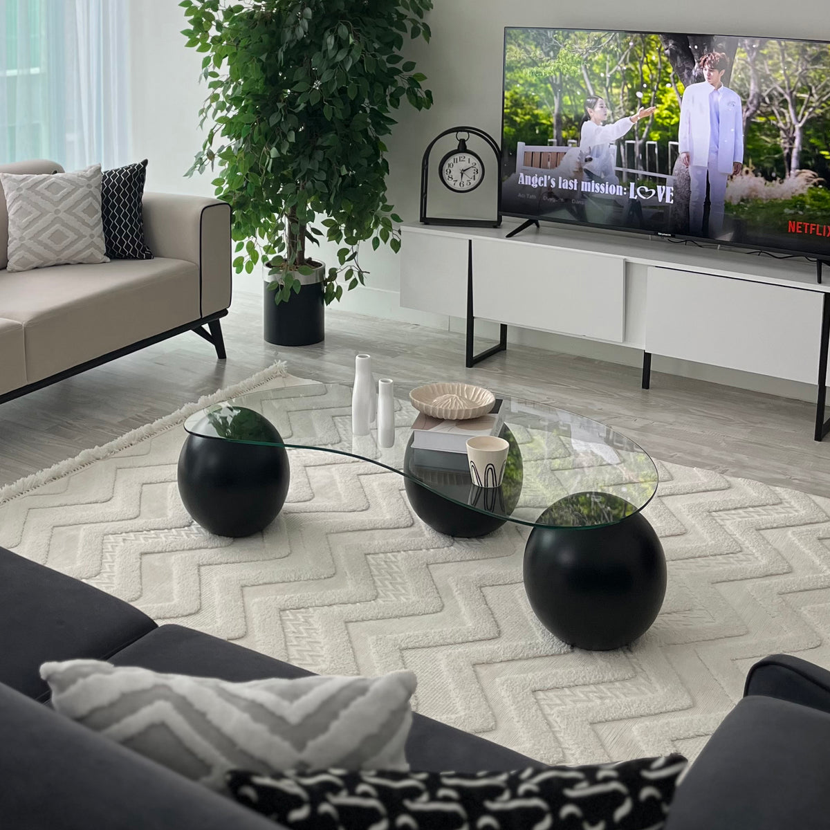 Wide living room view of the glass coffee table with black ball legs on a white textured rug before a TV unit