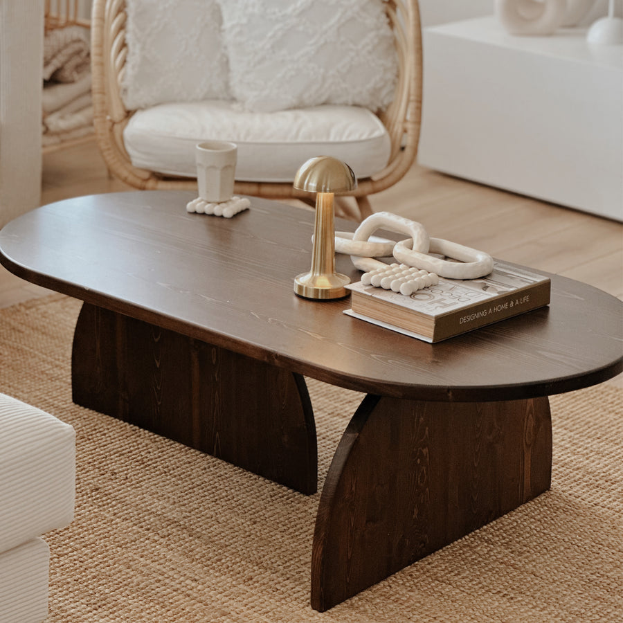 Dark brown oval Japandi coffee table of solid spruce with two half-round supports beside a sofa