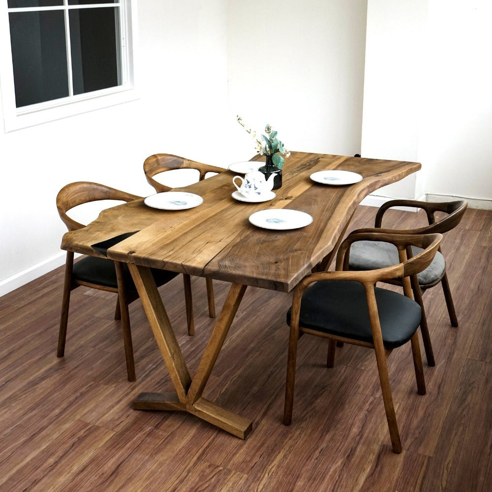 Walnut live edge farmhouse table with a V-shaped wooden base on a wood floor beside a window, corner view