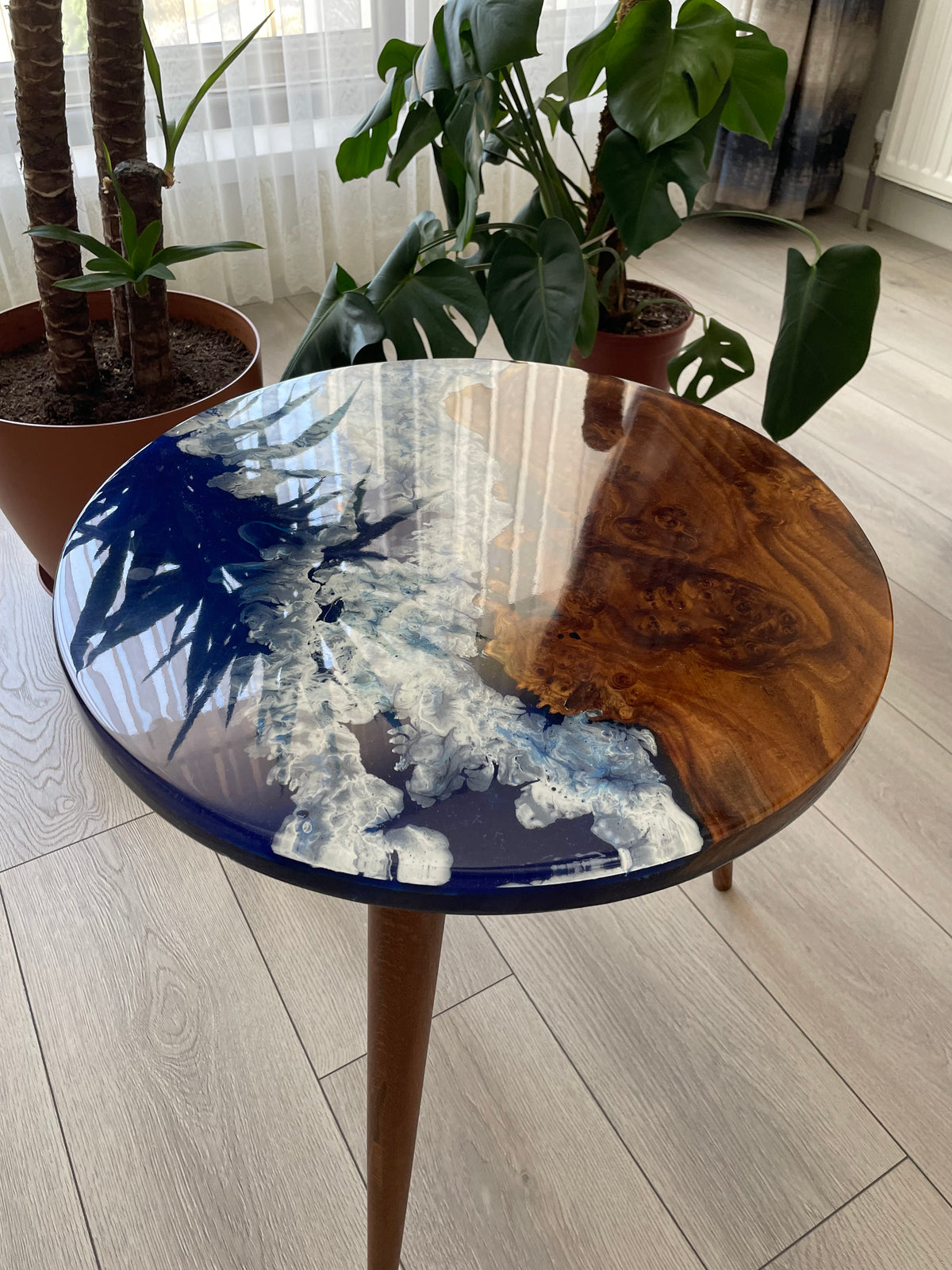 Round oleaster end table with a glossy ocean blue epoxy top and brown wood legs, with leaves reflected in the resin