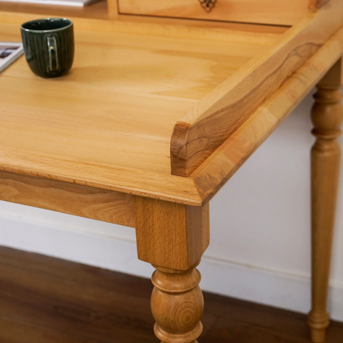 Close-up of the raised back edge and turned leg top of the beech writing desk, with a mug