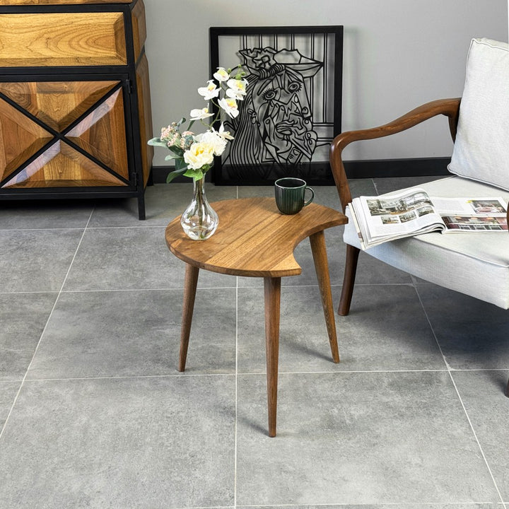 Distant view of the walnut C-shaped side table beside an armchair in front of a sideboard