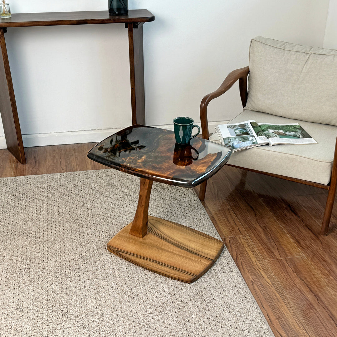 Dark brown walnut C shaped side table with a clear epoxy edge beside an armchair on a woven rug