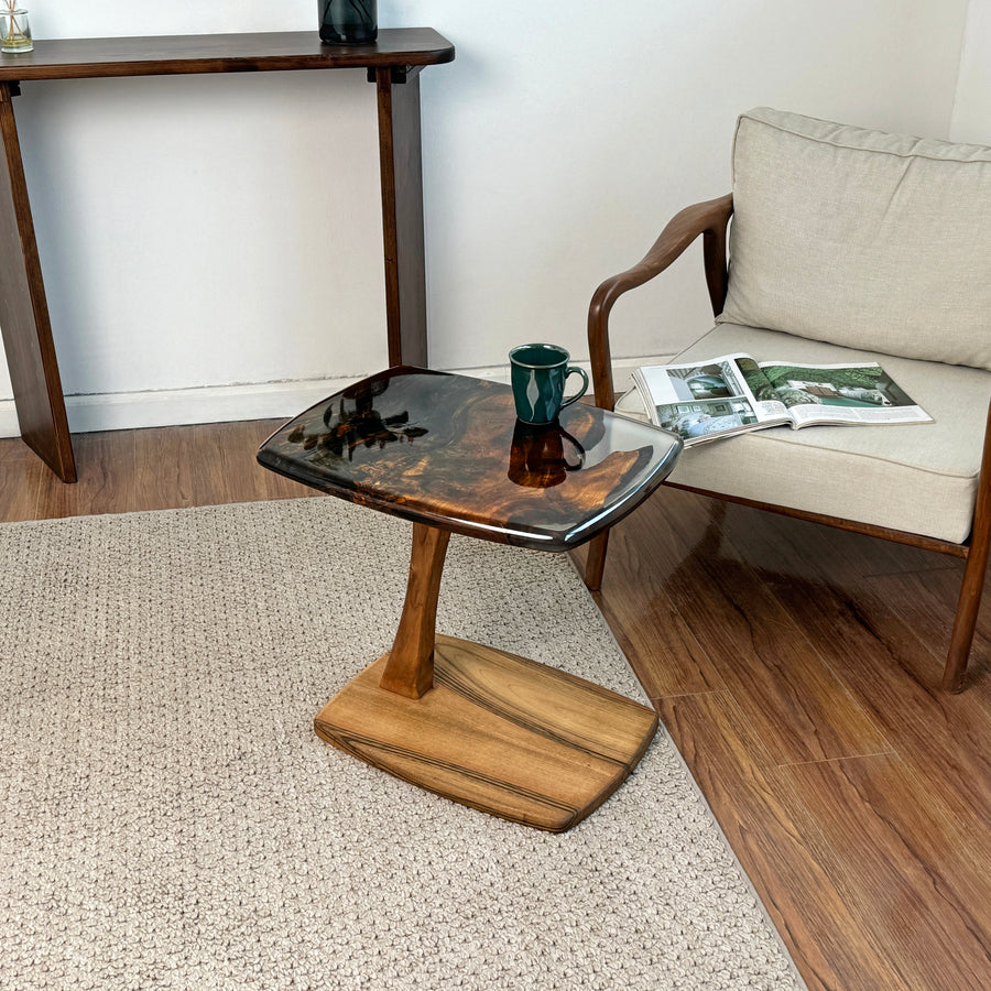 Dark brown walnut C shaped side table with a clear epoxy edge beside an armchair on a woven rug