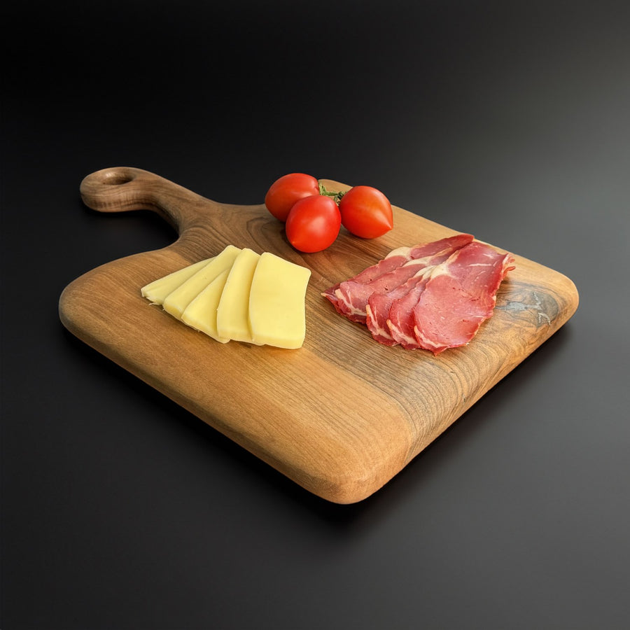 Walnut serving board with a looped handle at the back left, angled view on a black surface with tomatoes on top