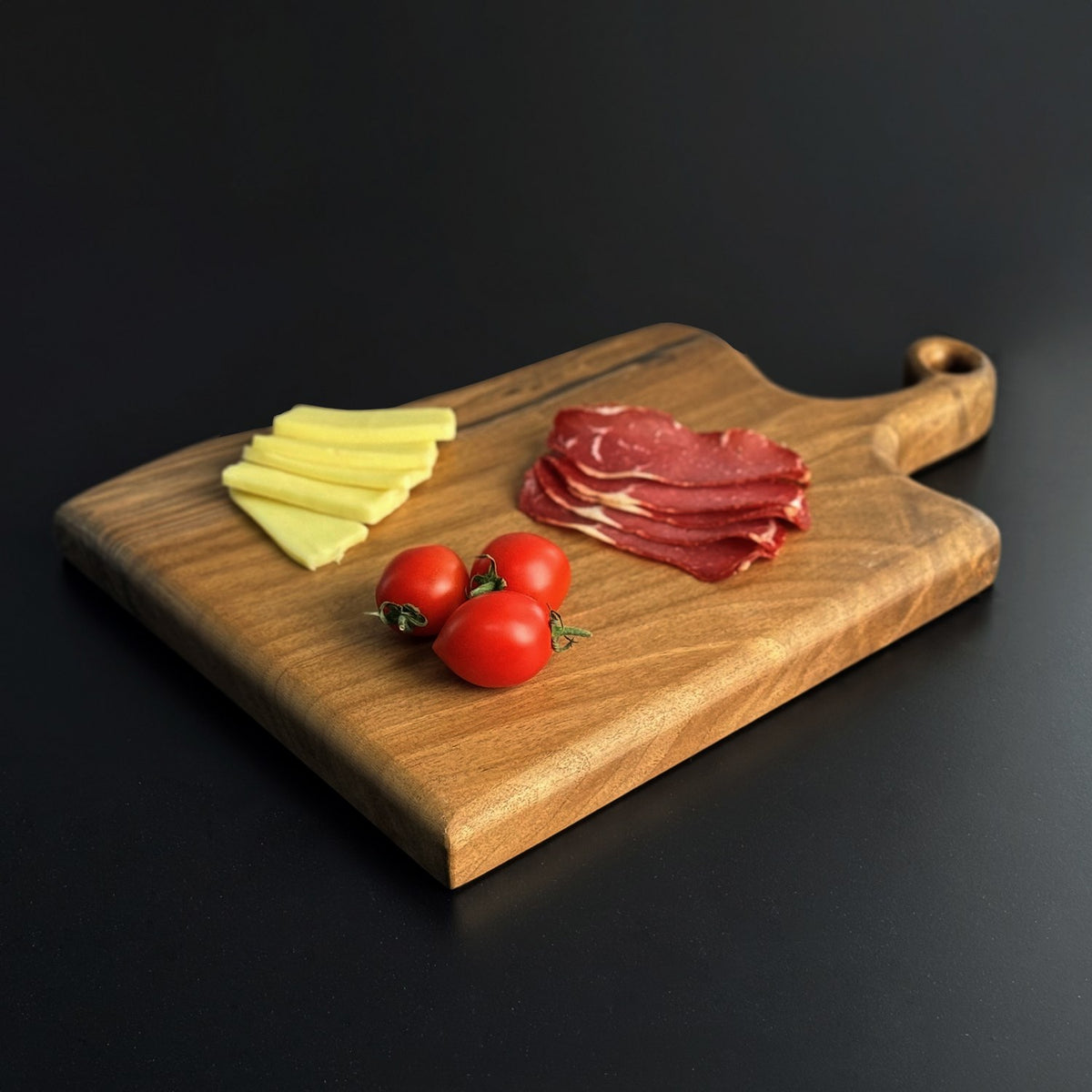 Walnut wood chopping board with a looped handle at the back right, angled view with sliced cheese on top