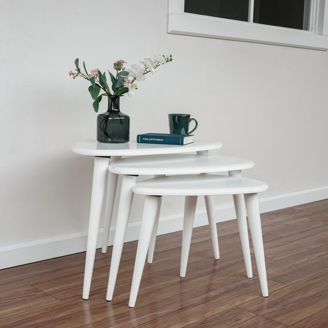 White nesting coffee table set of 3 on a wood floor in a room corner with a window, wide view