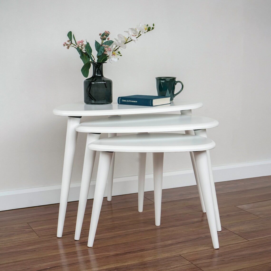 Low front view of the white nesting coffee table set of 3 with a vase, book and green mug on top