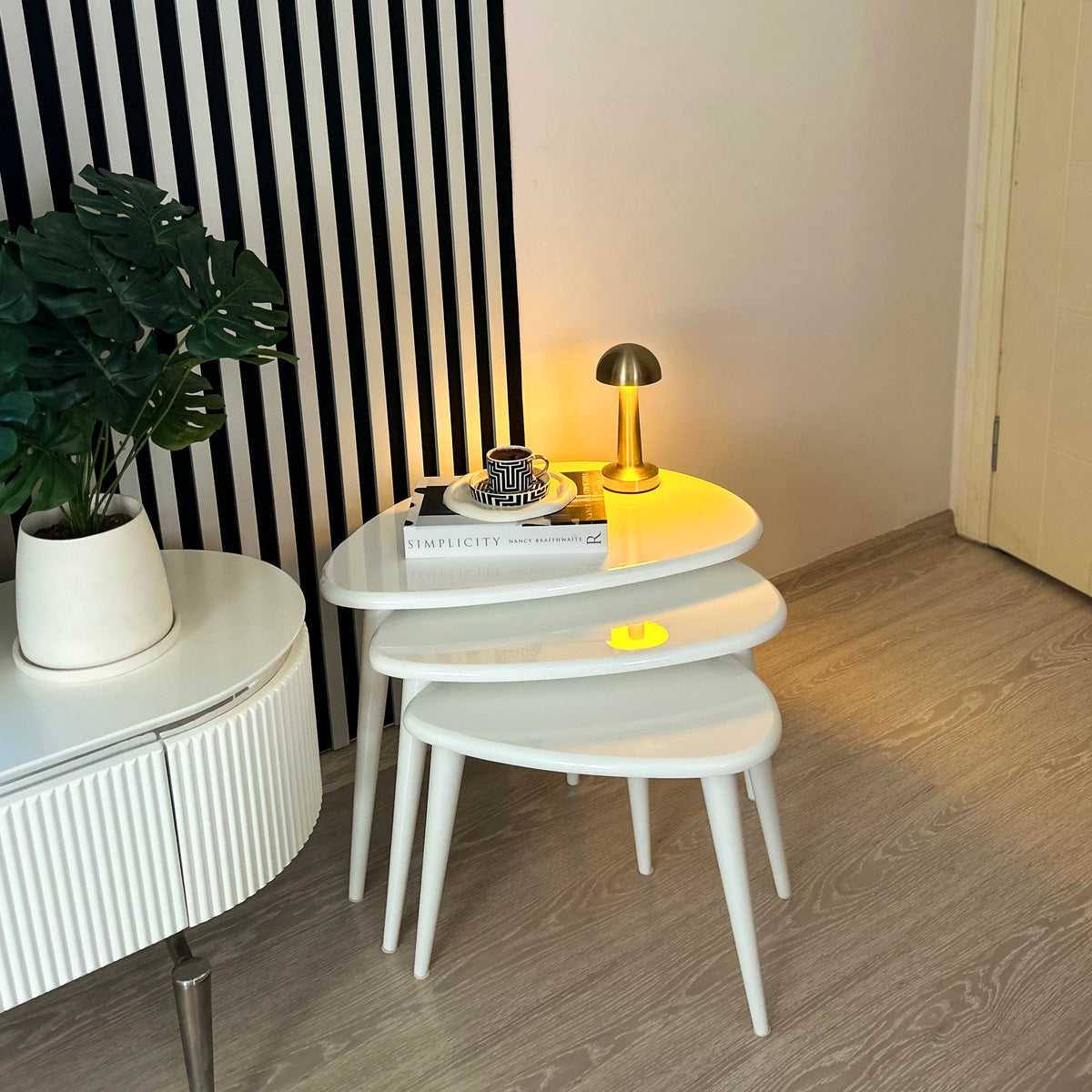 White high gloss nesting coffee table set of 3 with a gold lamp, in front of a striped wall