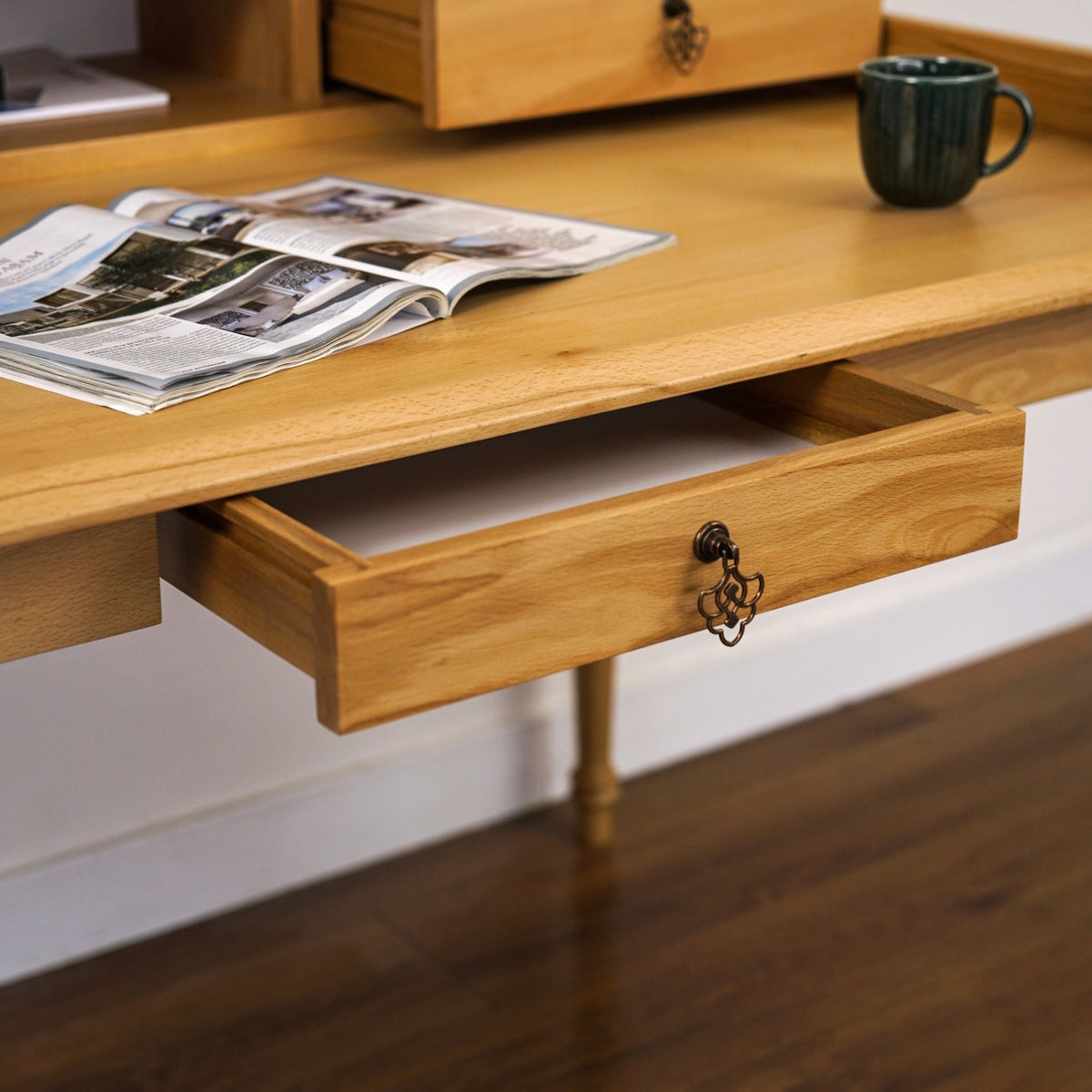 Close-up of the open center drawer of the beech writing desk with an antique-style pull and a mug