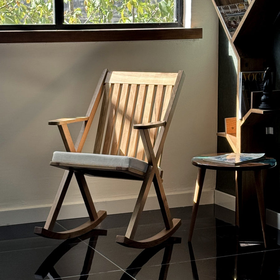 Solid wood walnut rocking chair with a gray seat cushion beside a round side table near a window