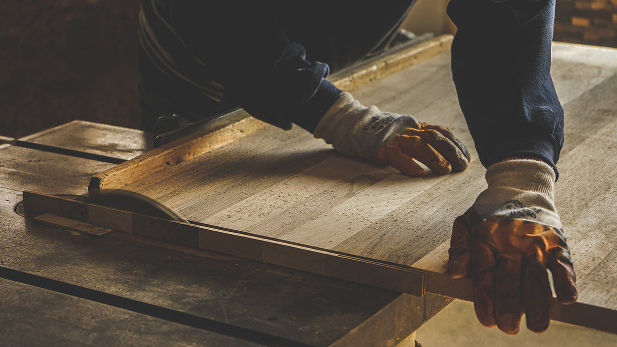 Woodworker in gloves guiding a wooden board across a table saw