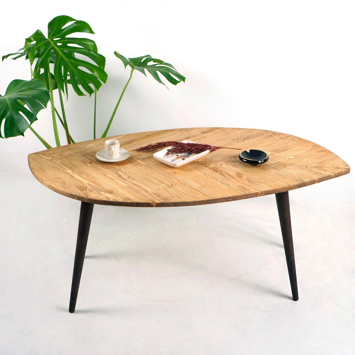 Leaf shaped solid wood coffee table with black tapered legs on a white background beside a monstera
